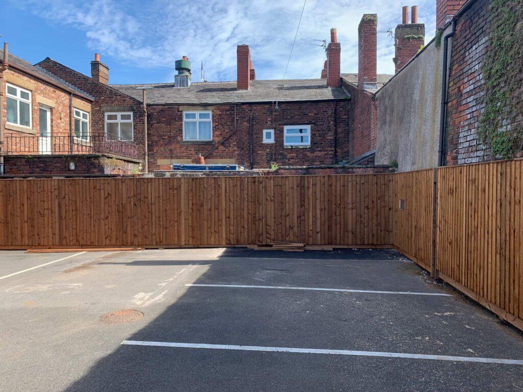 A parking lot with marked spaces is bordered by a tall wooden fence. Behind the fence, there are brick buildings with chimneys and windows. The sky is partly cloudy.