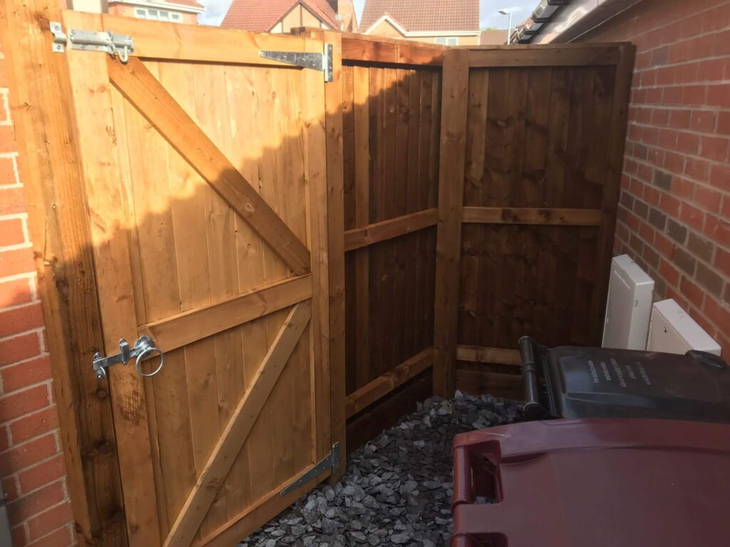 A wooden outdoor gate with a metal latch stands partially open, surrounded by a matching fence. The area is paved with dark gravel and contains a maroon wheelie bin. Brick walls and part of a house are visible in the background.