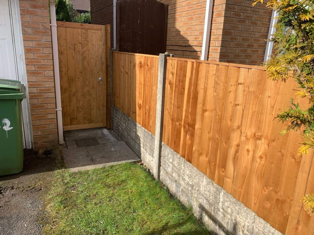 A narrow path leads to a wooden gate flanked by brick walls and a wooden fence. Theres a green trash bin on the left and a small patch of grass on the ground. The scene is brightly lit, indicating a sunny day.