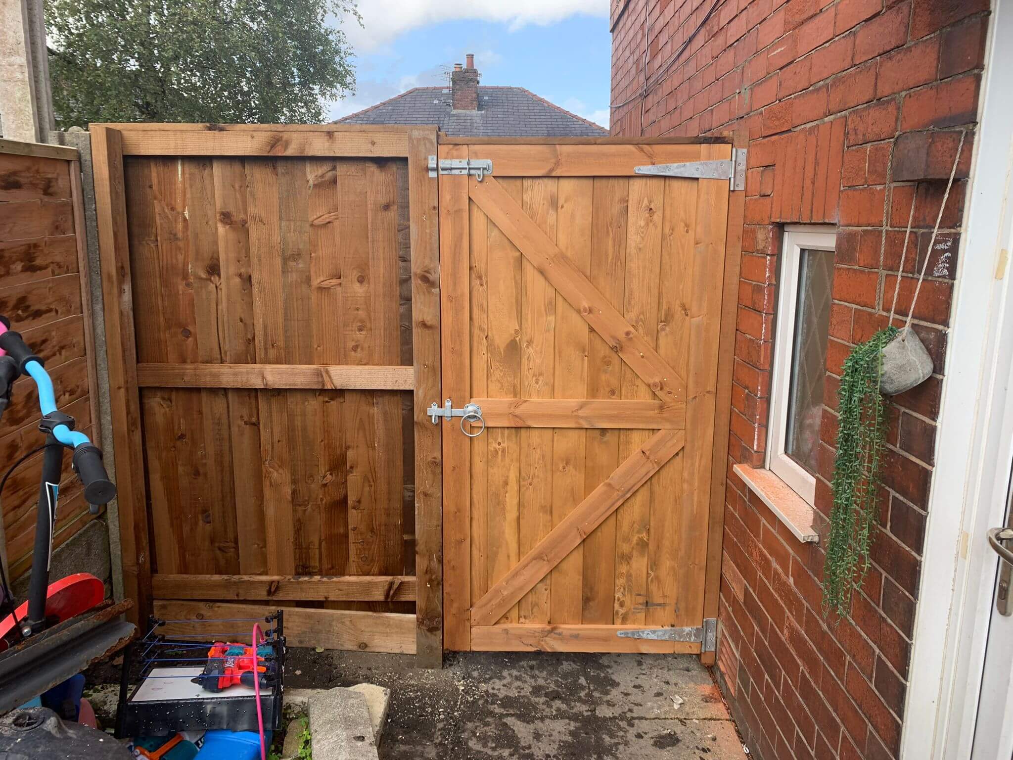 Wooden gate and fence in an alley beside a red brick house. The gate has a metallic latch. A small window is on the right with a hanging planter. A childs bicycle is partially visible on the left.