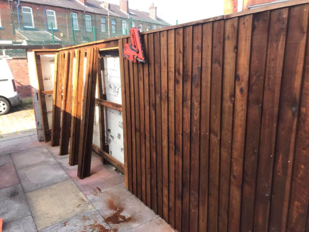 A neatly constructed dark wood fence under a clear sky, with several wooden panels leaning against it and a tool attached to the top. The fence stands on a paved area next to a row of brick buildings.