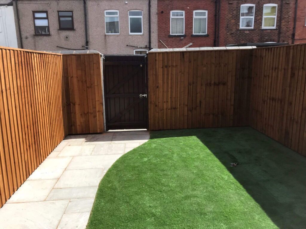 A small backyard with artificial grass and stone paving, surrounded by tall wooden fences. A wooden gate is partially open at the back, leading to a row of brick houses with visible windows.