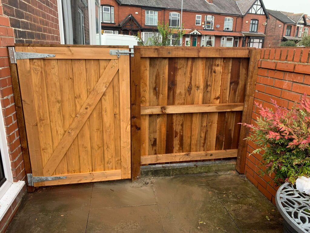 A wooden gate and fence are set in a brick wall on a stone patio. The gate has a diagonal support beam, and the fence has horizontal panels. In the background are brick houses with windows, trees, and a van parked on the street.