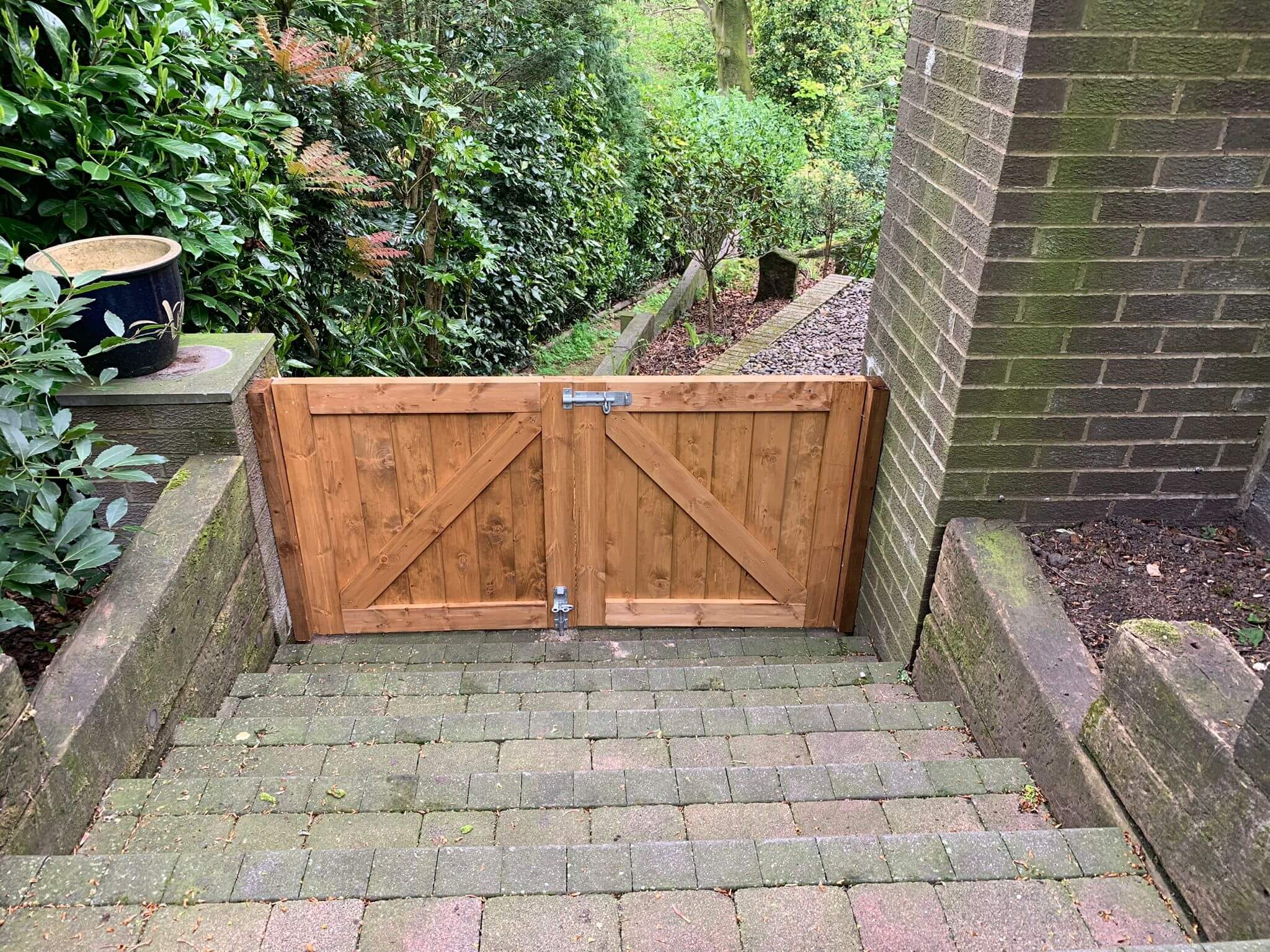 A wooden gate at the bottom of a stone staircase, surrounded by lush green foliage and brick walls on either side. The path beyond the gate leads into a garden area.